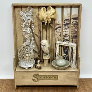 May include: A wooden shadow box with various vintage-style items. It features a small owl, a birdcage, a miniature duck, a small pitcher and bowl, a framed photo, and the text "Winton's". The box has a rustic, antique aesthetic.