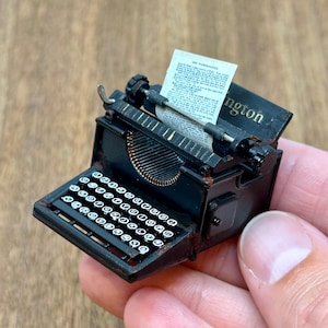 Puede incluir: Una máquina de escribir negra en miniatura con papel insertado, con un teclado con teclas blancas y una palanca de retorno de carro plateada. La palabra "Wellington" es visible en la parte superior. La máquina de escribir se sostiene en una mano.