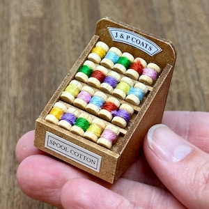 May include: A miniature wooden spool cotton display with a variety of coloured spools of thread. The display has a sign that reads "J&P Coats" and "Spool Cotton".