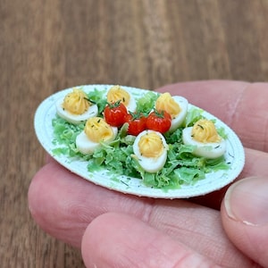 Pode incluir: Miniatura de ovos recheados e tomates cereja num prato branco com alface verde. Os ovos são cobertos com recheio amarelo e uma pitada de ervas. Um pequeno item de comida, perfeito para uma casa de bonecas ou uma coleção em miniatura.