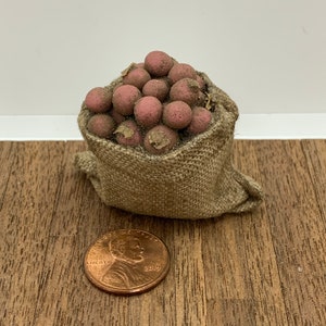 May include: A burlap sack filled with a large quantity of red potatoes. The sack is sitting on a wooden surface next to a US penny.