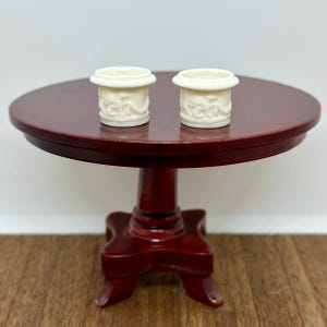 May include: Two white ceramic planters with an embossed floral design, sitting on a dark red wooden table.