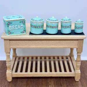 Puede incluir: Una panera miniatura de color azul claro y cuatro botes a juego con motivos florales y tapas, expuestos sobre una pequeña mesa de madera con estante de listones. La panera lleva la palabra BREAD en letras blancas en relieve al frente.