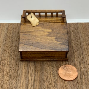 May include: A miniature wooden desk with a small railing on top. The desk is closed and has a brown stain finish.