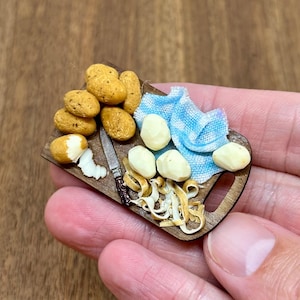 May include: Miniature wooden cutting board with potatoes, a small knife, and a blue and white checkered cloth. Some potatoes are peeled, and some are whole. The miniature food is a charming addition to a dollhouse or craft project.