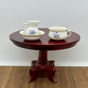 May include: A miniature white porcelain jug and chamber pot with blue floral designs, sitting on a dark brown wooden table.