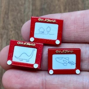 Puede incluir: Tres juguetes de dibujo en miniatura Etch A Sketch rojos con perillas blancas. Cada uno tiene una pantalla blanca con un dibujo de línea negra: una casa con árbol, un dinosaurio y un cohete. El nombre de la marca está impreso en dorado.