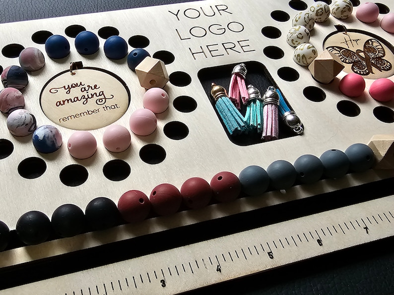 K&ouml;nnte beinhalten: Ein Holzperlenbrett mit verschiedenen Perlen in Rosa, Blau, Grau und Schwarz. Das Brett enth&auml;lt einen Bereich f&uuml;r Quasten und einen kreisf&ouml;rmigen Ausschnitt mit dem Text "you are amazing remember that."