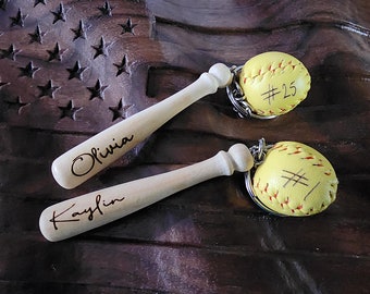 May include: Two softball-themed keychains. Each keychain features a small, yellow softball with red stitching and a wooden baseball bat. One softball has the number 25 written on it. The bats are engraved with the names "Olivia" and "Kaylin".
