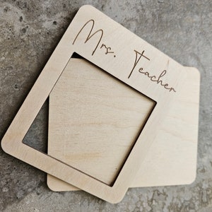 May include: Two light-colored wooden frames, one atop the other, with rounded corners. The top frame has the words "Mrs. Teacher" etched into the wood. The frames are designed to hold a photo or artwork.