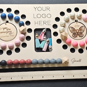 K&ouml;nnte beinhalten: Ein Holzbrett mit verschiedenen Perlen in Blau-, Rosa-, Grau- und Schwarzt&ouml;nen. Das Brett hat ein Lineal, den Text "YOUR LOGO HERE" und die W&ouml;rter "Small" und "Large". Es gibt auch Quasten und Holzformen.