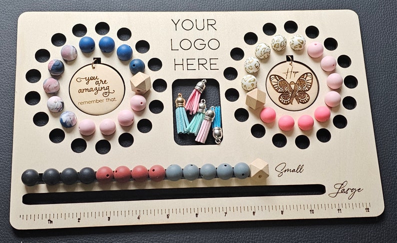 K&ouml;nnte beinhalten: Ein Holzbrett mit verschiedenen Perlen in Blau-, Rosa-, Grau- und Schwarzt&ouml;nen. Das Brett hat ein Lineal, den Text "YOUR LOGO HERE" und die W&ouml;rter "Small" und "Large". Es gibt auch Quasten und Holzformen.