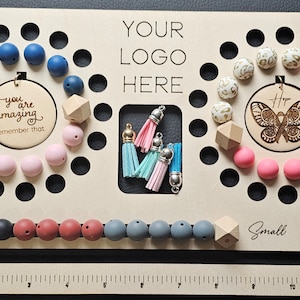 K&ouml;nnte beinhalten: Ein Holzperlenbrett mit verschiedenen Perlen in Blau-, Rosa- und Wei&szlig;t&ouml;nen. Das Brett hat einen Bereich f&uuml;r Quasten und enth&auml;lt den Text "YOUR LOGO HERE". Es gibt auch zwei Holzkreise mit Text und einem Schmetterlingsdesign.