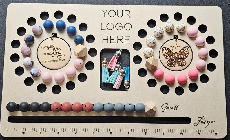 K&ouml;nnte beinhalten: Ein Holzperlenbrett mit verschiedenen Perlen in Blau-, Rosa- und Wei&szlig;t&ouml;nen. Das Brett hat einen Bereich f&uuml;r Quasten und enth&auml;lt den Text "YOUR LOGO HERE". Es gibt auch zwei Holzkreise mit Text und einem Schmetterlingsdesign.