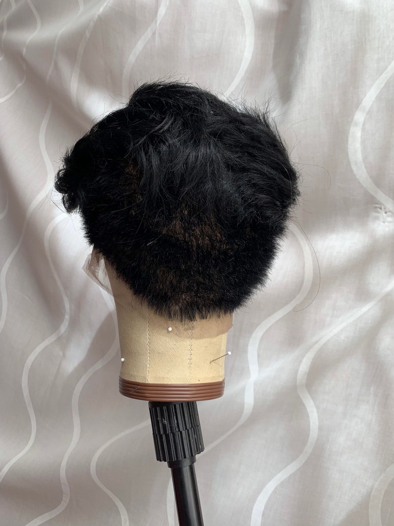 May include: Black wig on a mannequin head with a brown base and a black stand. The wig is styled in a short, textured look.