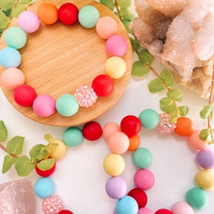 May include: Two colorful bracelets made of round beads. The bracelets are made of pastel colors, including pink, blue, green, yellow, and orange. Each bracelet has a single sparkly bead.