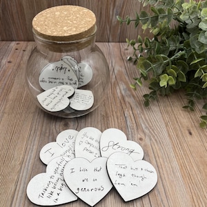 May include: A clear glass jar with a cork lid, filled with heart-shaped cards. Several cards are scattered around the jar. The cards have handwritten messages like "I love that you feel like being hugged in a big way."