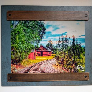 May include: A framed photograph of a red building with a train track leading away from it. The building is surrounded by trees and the sky is blue with white clouds. The frame is made of wood with a dark brown stain.