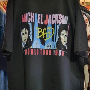 May include: A black short-sleeve t-shirt featuring a graphic print of Michael Jackson. The design includes the text "MICHAEL JACKSON", "BAD" in yellow, and "WORLD TOUR 1988" in red, alongside two portraits of the artist and blue silhouettes.