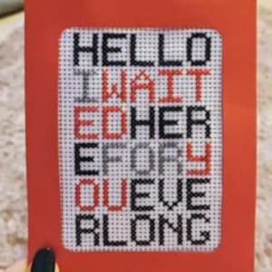 May include: A cross-stitch card with the words "HELLO I WAIT ED HER E DRY QUEUE R LONG" in black and red thread. The card is set in a red frame.