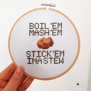 Könnte beinhalten: Ein weißer Stickrahmen mit dem Text "BOIL 'EM MASH 'EM STICK 'EM IN A STEW" und einem braunen Kartoffelbild.