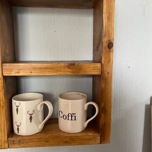 May include: Two white ceramic mugs on a wooden shelf. One mug has a deer pattern and the other mug says "Coffe" in black lettering.