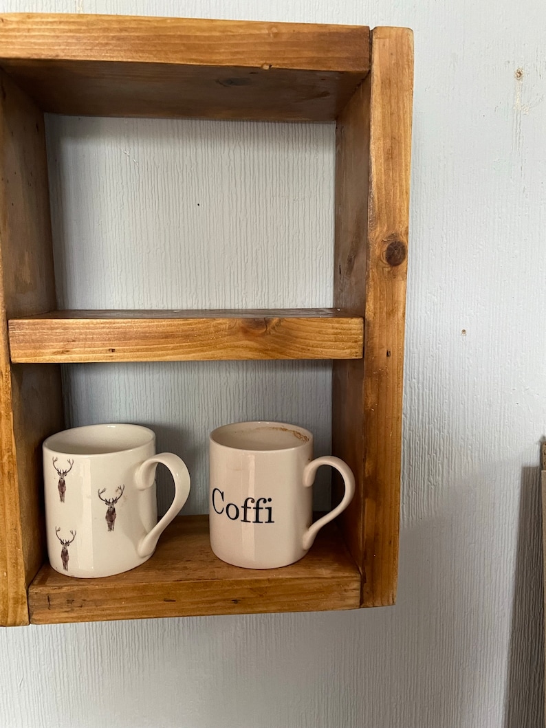 May include: Two white ceramic mugs on a wooden shelf. One mug has a deer pattern and the other mug says "Coffe" in black lettering.