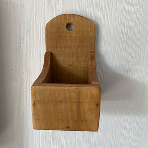 Pu&ograve; includere: Un piccolo contenitore rettangolare in legno da parete con la parte superiore curva e un foro per appenderlo. Il contenitore &egrave; realizzato in legno chiaro e ha un design semplice e rustico. Il contenitore &egrave; vuoto.