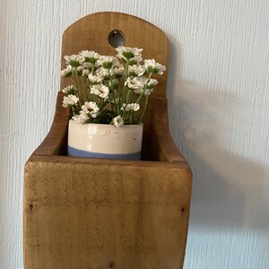 Pu&ograve; includere: Un'antica fioriera in legno da parete con finitura anticata. Contiene un piccolo vaso in ceramica bianco e blu pieno di fiori bianchi simili a margherite. La fioriera ha una forma a scatola rettangolare e una parte superiore arrotondata con un foro per appenderla.