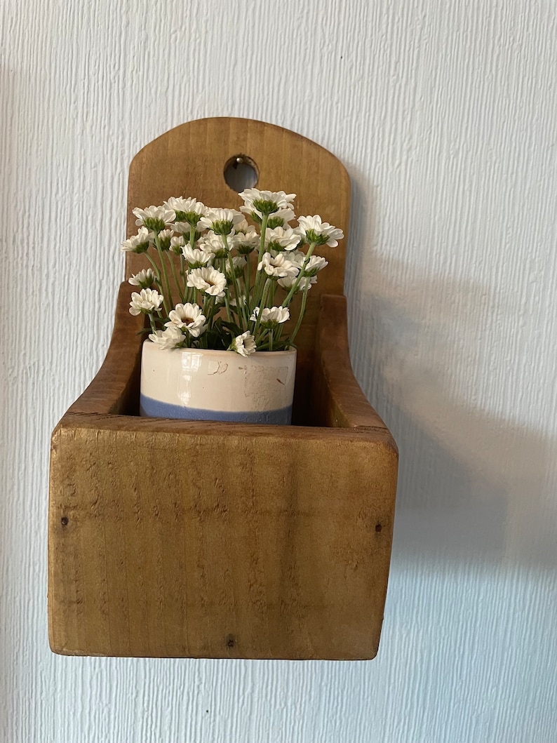 Pu&ograve; includere: Un'antica fioriera in legno da parete con finitura anticata. Contiene un piccolo vaso in ceramica bianco e blu pieno di fiori bianchi simili a margherite. La fioriera ha una forma a scatola rettangolare e una parte superiore arrotondata con un foro per appenderla.