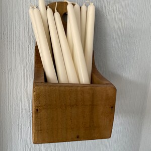 Pu&ograve; includere: Un portacandele in legno pieno di candele affusolate color avorio. Il supporto ha una forma rettangolare rustica con una parte superiore curva ed &egrave; montato su una parete azzurra. Le candele sono disposte verticalmente, creando un'esposizione semplice ed elegante.