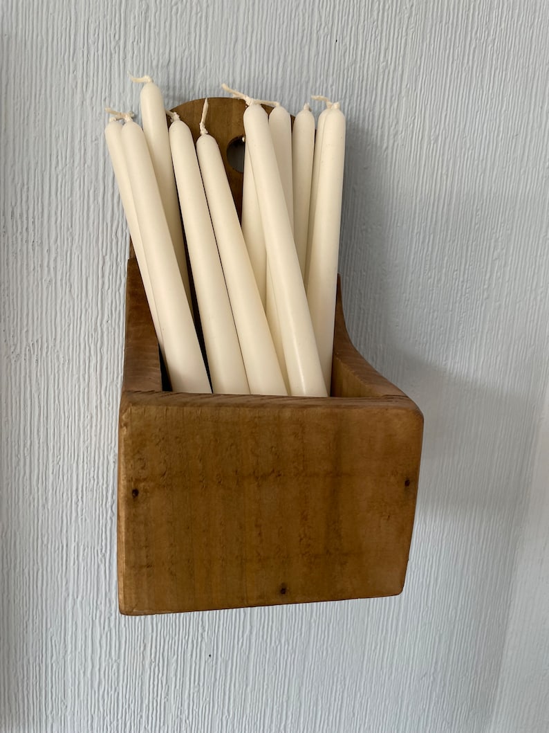 Pu&ograve; includere: Un portacandele in legno pieno di candele affusolate color avorio. Il supporto ha una forma rettangolare rustica con una parte superiore curva ed &egrave; montato su una parete azzurra. Le candele sono disposte verticalmente, creando un'esposizione semplice ed elegante.