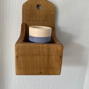 Pu&ograve; includere: Un portapezie rustico in legno da parete con un piccolo contenitore in ceramica bicolore all'interno. Il contenitore &egrave; azzurro nella parte inferiore e color crema nella parte superiore. Il portapezie &egrave; in legno non rifinito e ha un foro nella parte superiore.