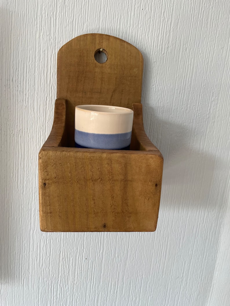 Pu&ograve; includere: Un portapezie rustico in legno da parete con un piccolo contenitore in ceramica bicolore all'interno. Il contenitore &egrave; azzurro nella parte inferiore e color crema nella parte superiore. Il portapezie &egrave; in legno non rifinito e ha un foro nella parte superiore.