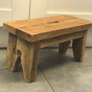 May include: A small, rustic wooden step stool. The stool is made of light brown, weathered wood with visible grain and knots. It has a rectangular top with a handhold and four sturdy legs. The stool measures approximately 46 cm long, 23 cm wide, and 20 cm tall.