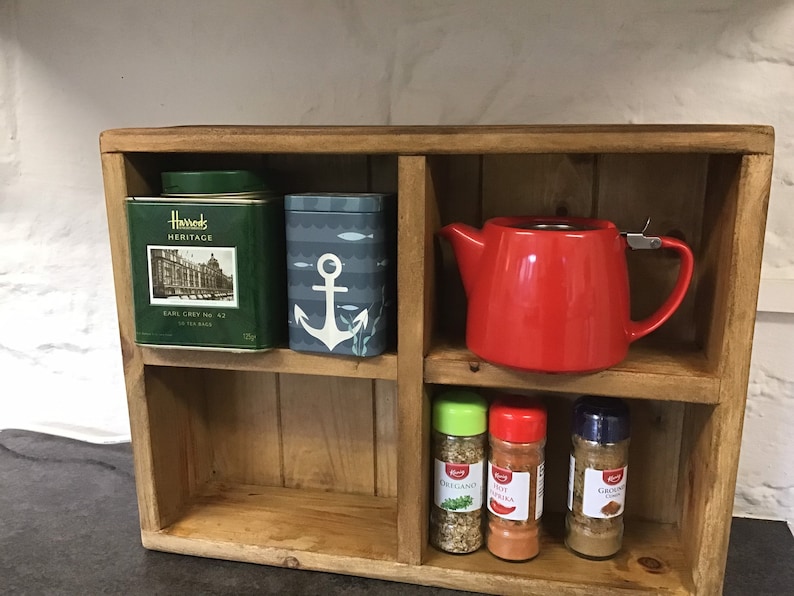 May include: A wooden shelf unit with four compartments. The top shelf has a green tea tin that says "Harrods Heritage Earl Grey No. 42" and a blue tin with an anchor design. The middle shelf has a red teapot. The bottom shelf has three spice jars with labels that say "Oregano", "Pote Paprika", and "Ground Cinnamon".