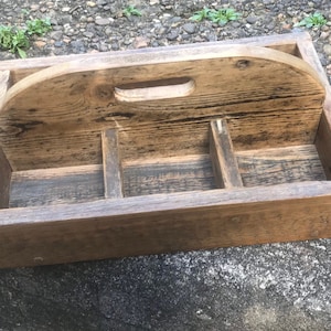 May include: A rectangular wooden caddy with a handle. The caddy is made of weathered wood with a natural finish. It has a handle on top and dividers inside, suitable for organizing items. The caddy is designed for carrying or storing items.