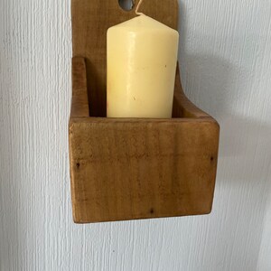Pu&ograve; includere: Un portacandele rustico in legno con finitura naturale, con una candela cilindrica color crema. Il supporto ha una base rettangolare e una parte superiore curva con un foro per appenderlo. La candela &egrave; alta circa 7.6 cm.