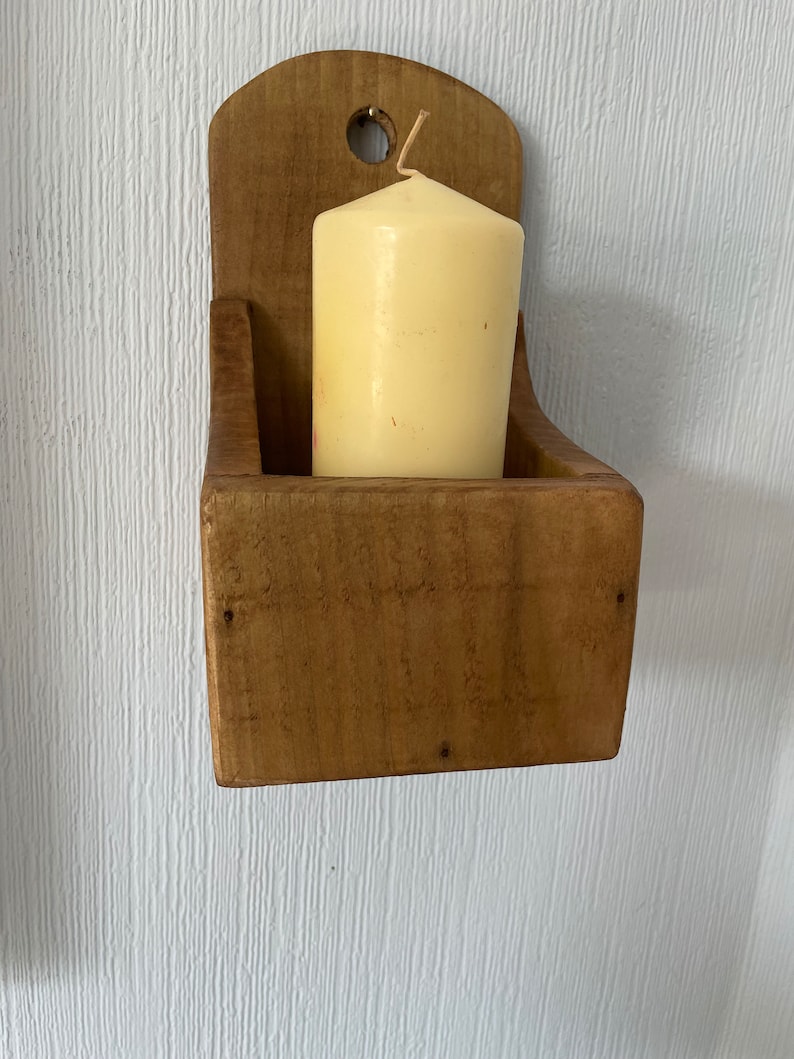 Pu&ograve; includere: Un portacandele rustico in legno con finitura naturale, con una candela cilindrica color crema. Il supporto ha una base rettangolare e una parte superiore curva con un foro per appenderlo. La candela &egrave; alta circa 7.6 cm.