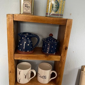 May include: A wooden shelf with two shelves, displaying various items. The top shelf has two tin canisters, one with the text "Afternoon Tea" and the other with the text "Harrods". The middle shelf has a blue and white polka dot pitcher and a blue and white polka dot lidded jar. The bottom shelf has two white mugs, one with a reindeer design and the other with the text "Coffe".