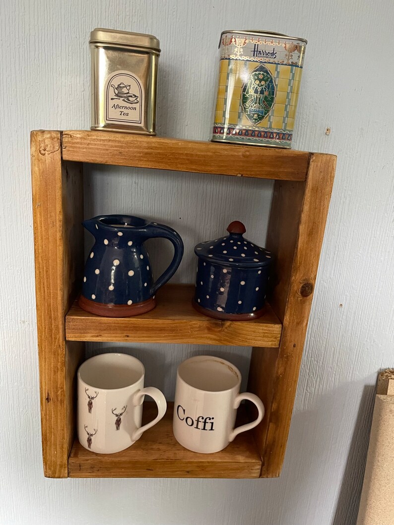 May include: A wooden shelf with two shelves, displaying various items. The top shelf has two tin canisters, one with the text "Afternoon Tea" and the other with the text "Harrods". The middle shelf has a blue and white polka dot pitcher and a blue and white polka dot lidded jar. The bottom shelf has two white mugs, one with a reindeer design and the other with the text "Coffe".