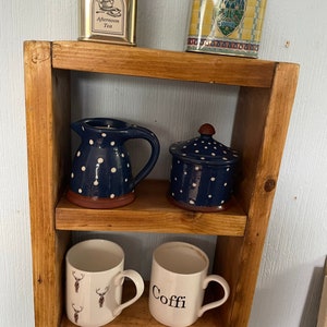May include: A wooden shelf with three shelves. The top shelf has two tin boxes, one with the text "Harrods" and the other with the text "Afternoon Tea". The middle shelf has a blue and white polka dot pitcher and a blue and white polka dot lidded jar. The bottom shelf has two mugs, one with a deer pattern and the other with the text "Coffe".