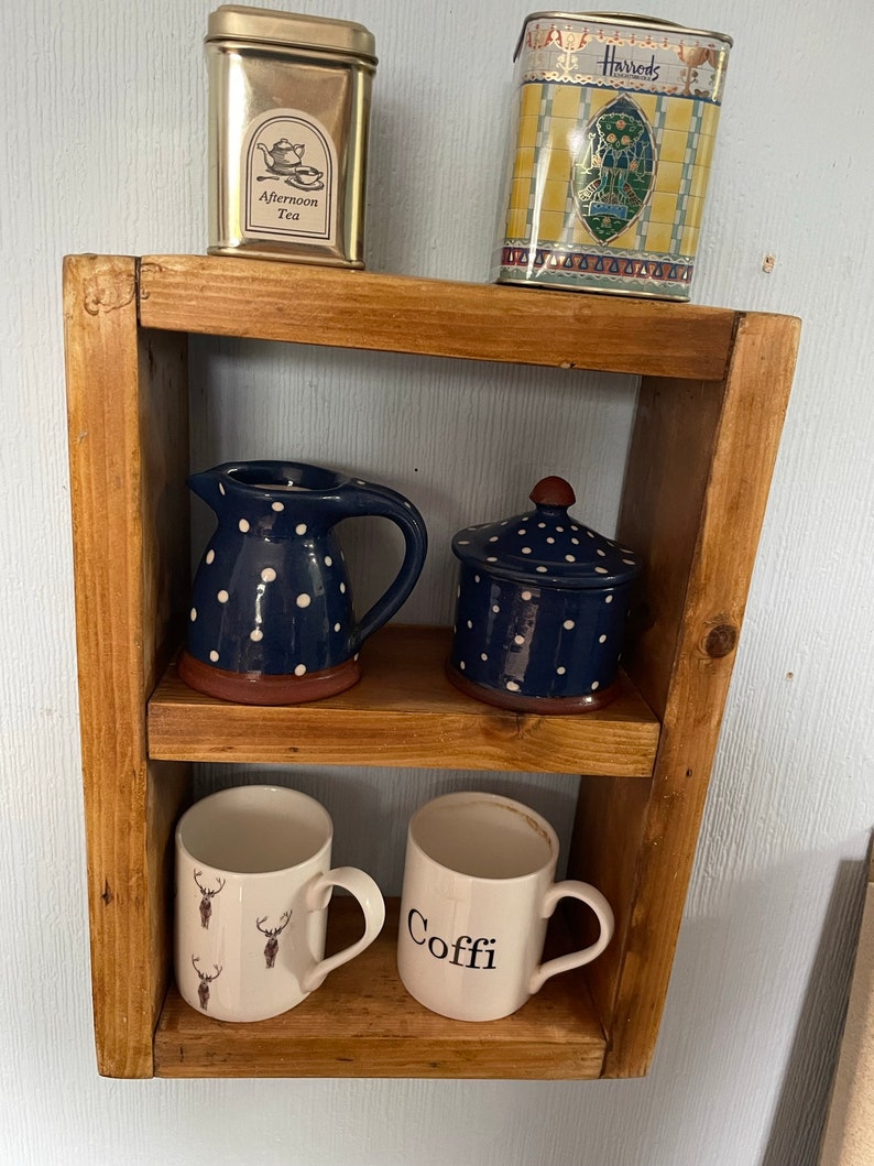 May include: A wooden shelf with three shelves. The top shelf has two tin boxes, one with the text "Harrods" and the other with the text "Afternoon Tea". The middle shelf has a blue and white polka dot pitcher and a blue and white polka dot lidded jar. The bottom shelf has two mugs, one with a deer pattern and the other with the text "Coffe".