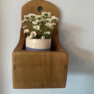 Pu&ograve; includere: Un'antica fioriera in legno montata a parete con la parte superiore curva e un piccolo foro. All'interno, un piccolo vaso di ceramica con una striscia blu contiene fiori bianchi. La fioriera ha una finitura in legno naturale.