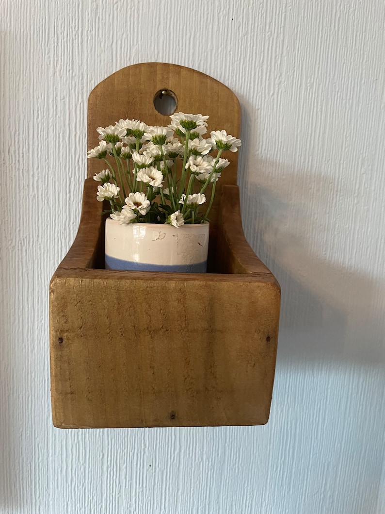 Pu&ograve; includere: Un'antica fioriera in legno montata a parete con la parte superiore curva e un piccolo foro. All'interno, un piccolo vaso di ceramica con una striscia blu contiene fiori bianchi. La fioriera ha una finitura in legno naturale.