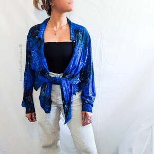 Könnte beinhalten: Ein blau gemustertes Hemd mit Knöpfen, das in der Taille gebunden ist, kombiniert mit einer weißen strukturierten Hose und einem schwarzen Bustier. Das Hemd hat ein lebendiges blaues und schwarzes Design. Das Outfit ist vor einem weißen Hintergrund.