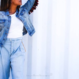 May include: A woman wearing a light blue denim jacket with fringe detail on the sleeves. The jacket has a white embroidered design on the left side. She is also wearing light blue denim pants with a white t-shirt underneath.