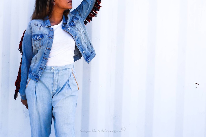 May include: A woman wearing a light blue denim jacket with fringe detail on the sleeves. The jacket has a white embroidered design on the left side. She is also wearing light blue denim pants with a white t-shirt underneath.