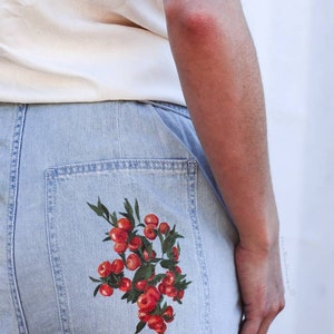 May include: Light blue denim jeans with a red and green painted design of a cluster of berries on the back pocket.
