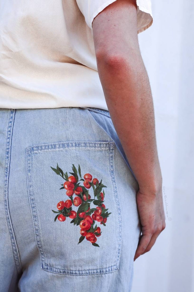May include: Light blue denim jeans with a red and green painted design of a cluster of berries on the back pocket.